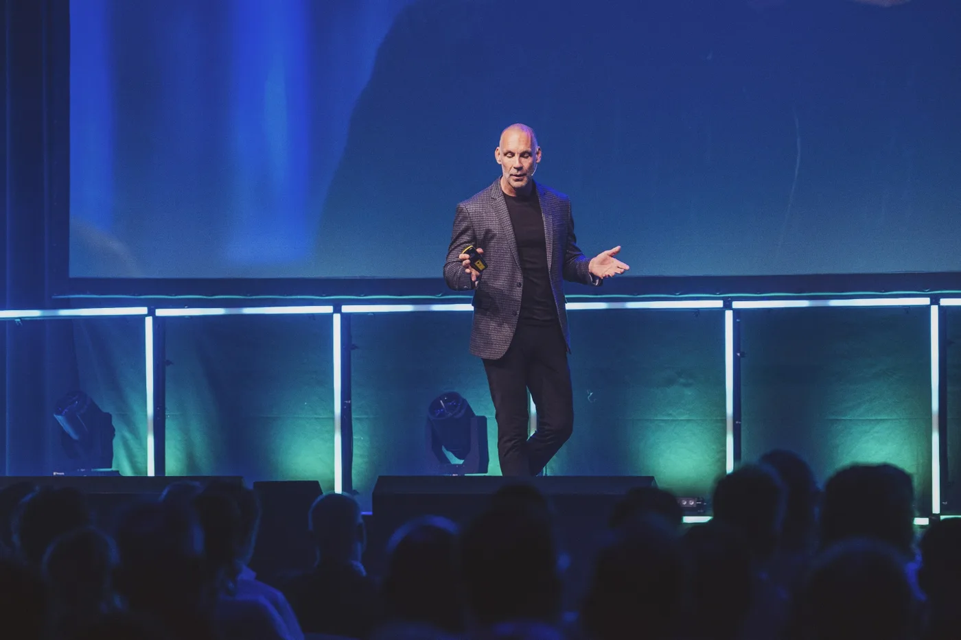 Richard van Hooijdonk in geruit colbert op een podium in blauwgroen licht, met het publiek als silhouet op de voorgrond