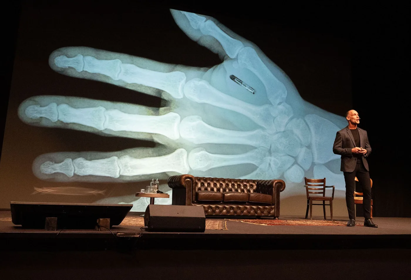 Richard op een theaterpodium naast een grote röntgenfoto van een hand met een chip