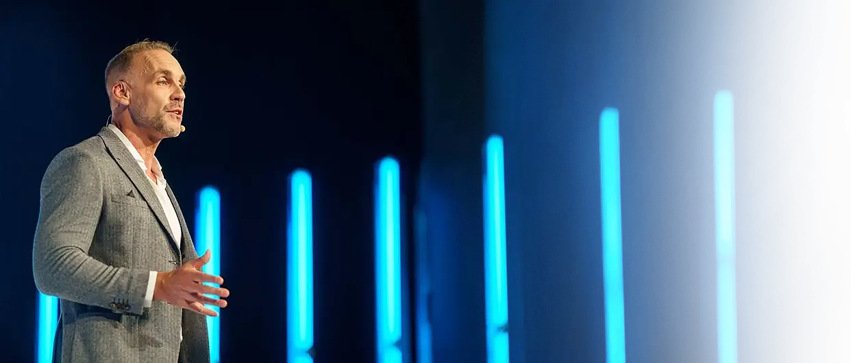 Richard van Hooijdonk in grijs colbert op het podium, voor blauwe verticale lichtlijnen